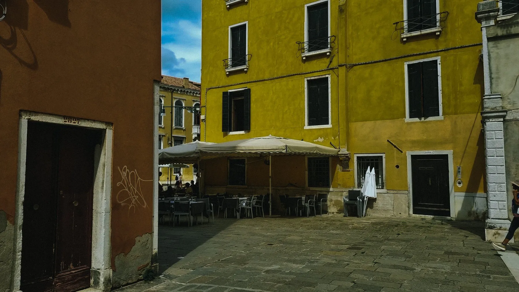 Café tables and a large parasol in a sunlit courtyard of ochre and yellow houses
