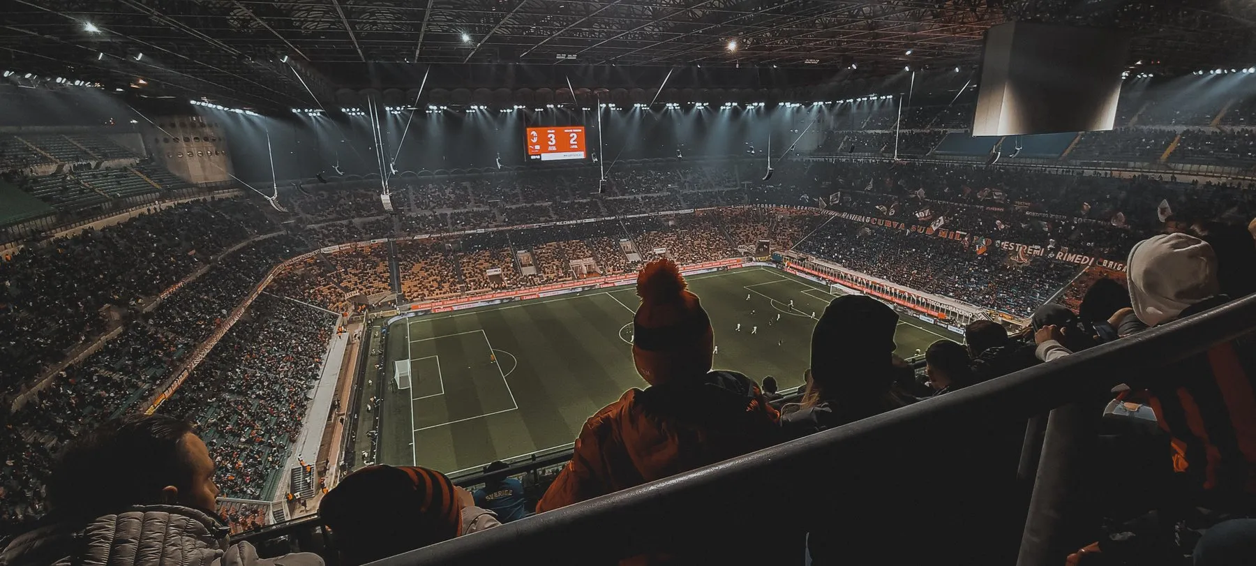 Floodlit night football match viewed from the upper stands, crowd around the pitch